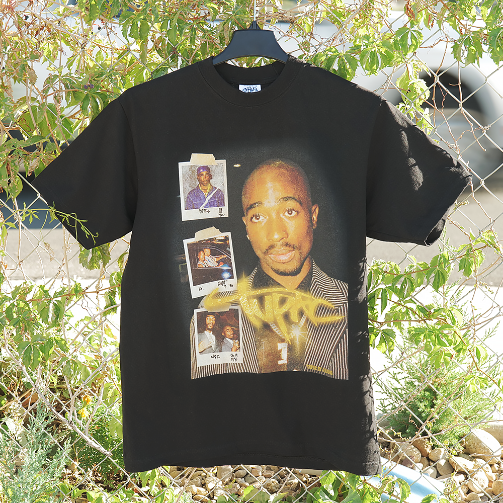 Black t-shirt with graphic design of a Tupac and polaroid photos, hanging against a natural background.