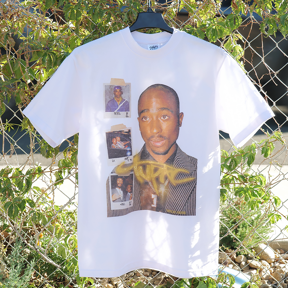 White t-shirt with graphic design of a Tupac and polaroid photos, hanging against a natural background.